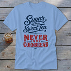 Light blue t-shirt featuring the text Sugar in my Sweat Tea NEVER IN MY CORNBREAD on a wooden surface.