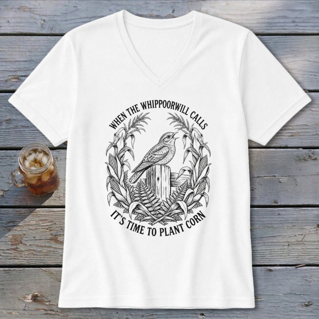 A white V-neck t-shirt with a black line art graphic of a bird on a fence post surrounded by corn stalks and the text "When the whippoorwill calls, it's time to plant corn."