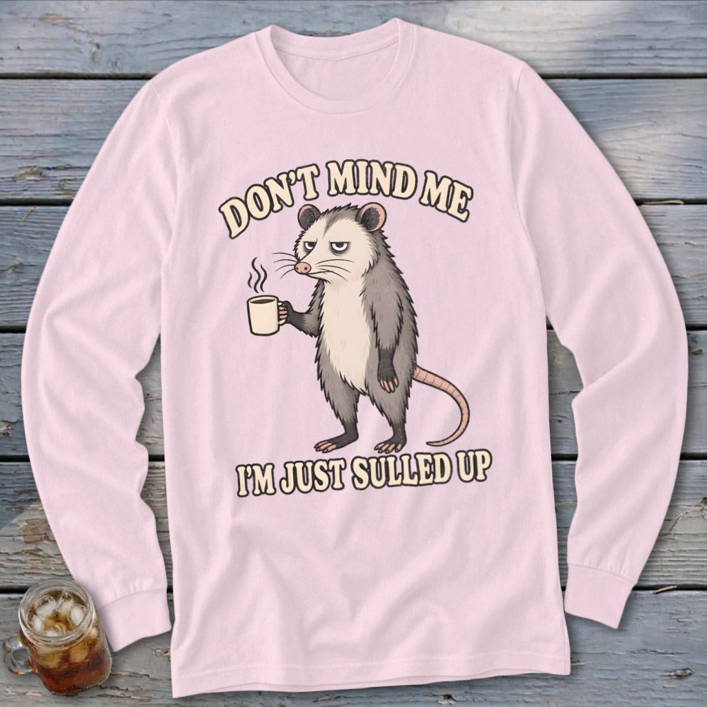 Pink long sleeve shirt with a grumpy opossum holding a coffee cup and text that reads "Don't mind me I'm just sulled up."