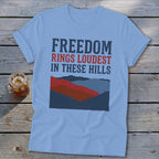 Light blue t-shirt featuring distressed text reading FREEDOM RINGS LOUDEST IN THESE HILLS above a stylized mountain graphic, laid on wooden boards next to a glass of iced tea.