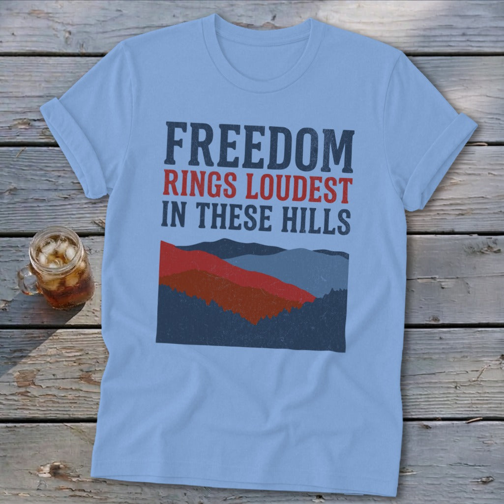 Light blue t-shirt featuring distressed text reading FREEDOM RINGS LOUDEST IN THESE HILLS above a stylized mountain graphic, laid on wooden boards next to a glass of iced tea.