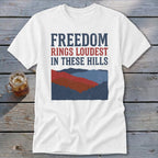 White t-shirt with red, white, and blue text that reads Freedom Rings Loudest In These Hills above a mountain landscape graphic.