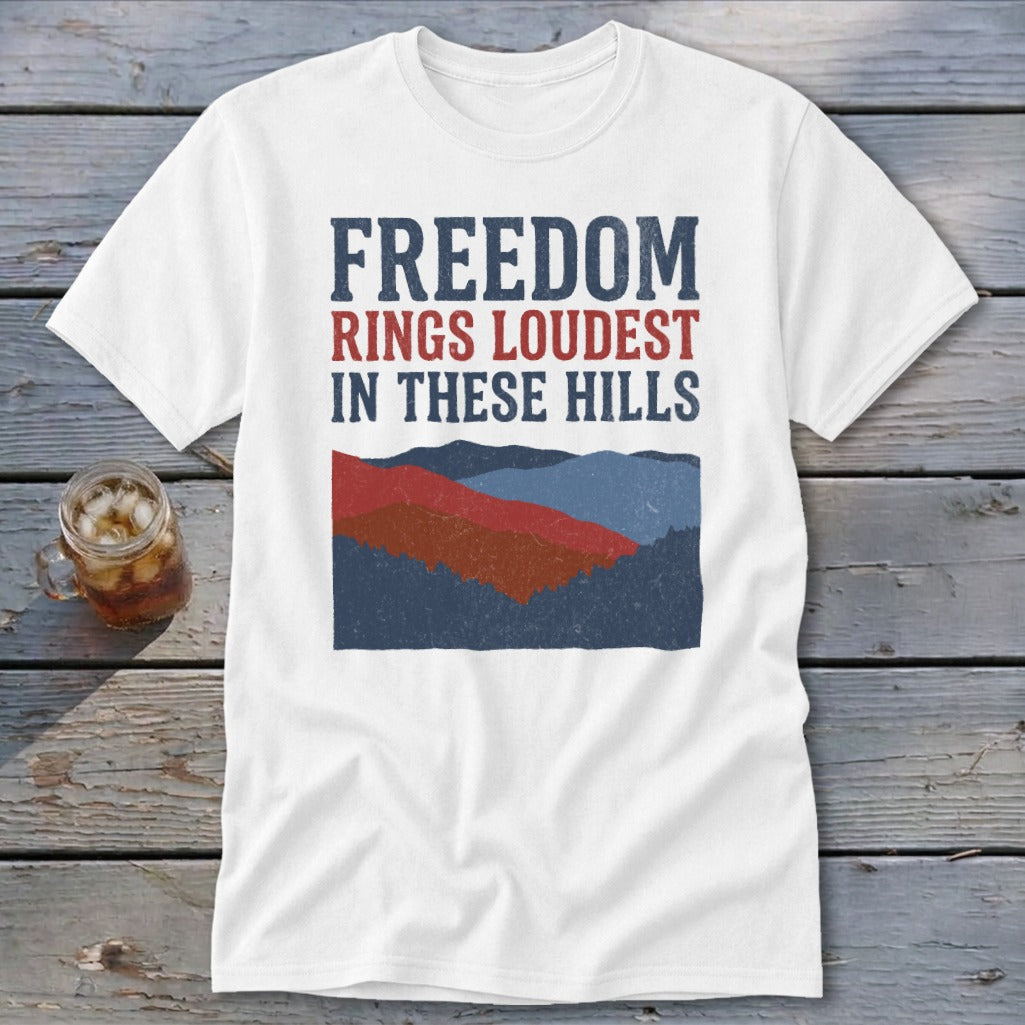 White t-shirt with red, white, and blue text that reads Freedom Rings Loudest In These Hills above a mountain landscape graphic.