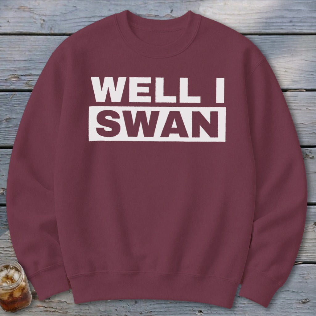 Maroon crewneck sweatshirt featuring white text that reads WELL I SWAN laid on a wooden background.