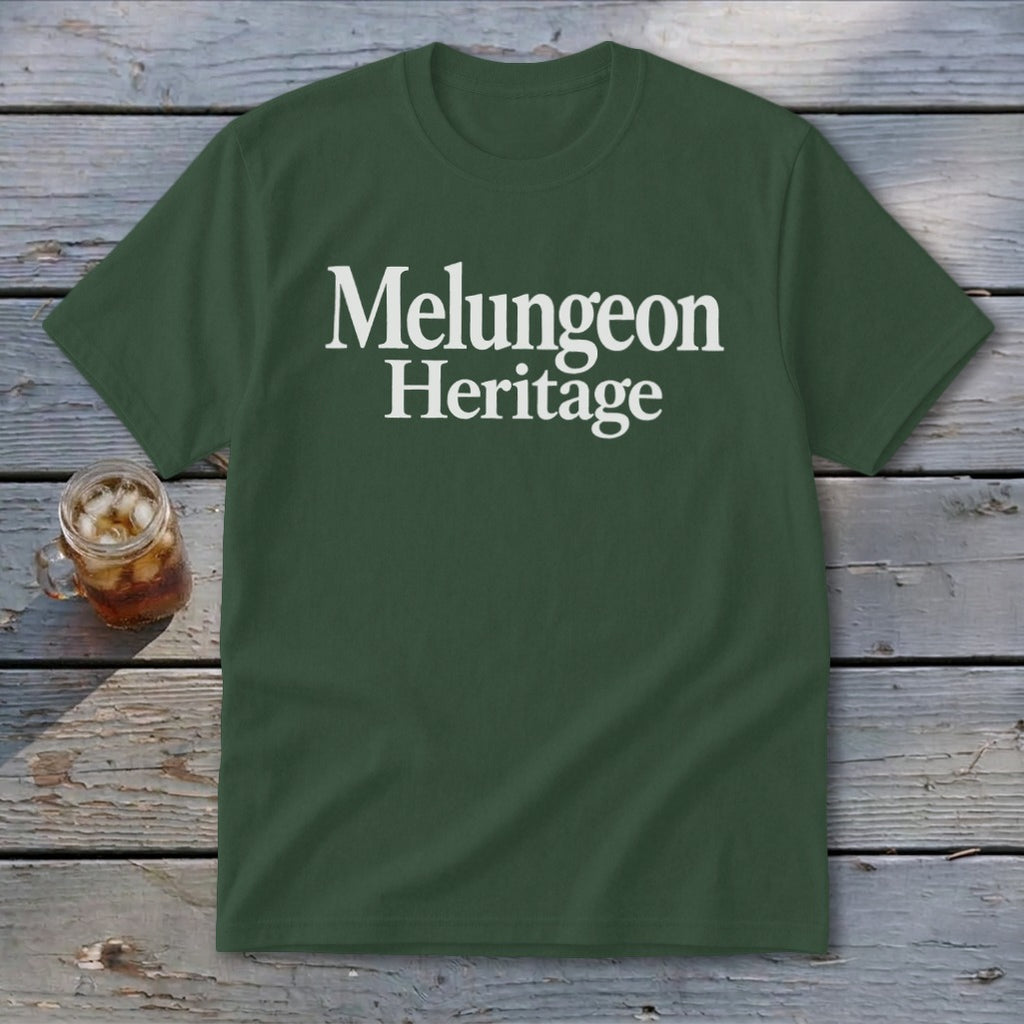 Forest green crewneck t-shirt with the white text "Melungeon Heritage" centered on the chest, laid flat on a rustic wooden surface next to a glass of iced tea.