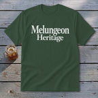 Forest green crewneck t-shirt with the white text "Melungeon Heritage" centered on the chest, laid flat on a rustic wooden surface next to a glass of iced tea.