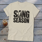 Cream t-shirt featuring distressed black text that reads SANG SEASON, displayed on a wooden surface with a glass beverage.