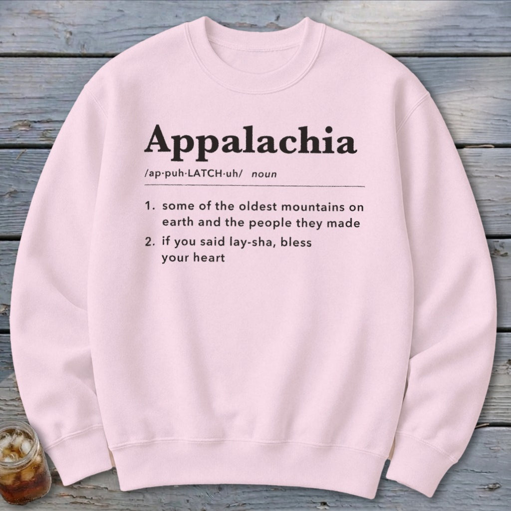 Light pink crewneck sweatshirt featuring black dictionary-style text defining Appalachia displayed centrally against a wooden background.