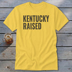 Yellow t-shirt with black text reading "KENTUCKY RAISED" displayed on wooden planks.