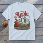 A light blue t-shirt printed with a vintage graphic of an apple, cabin, and mountains, labeled Apple At Cha.