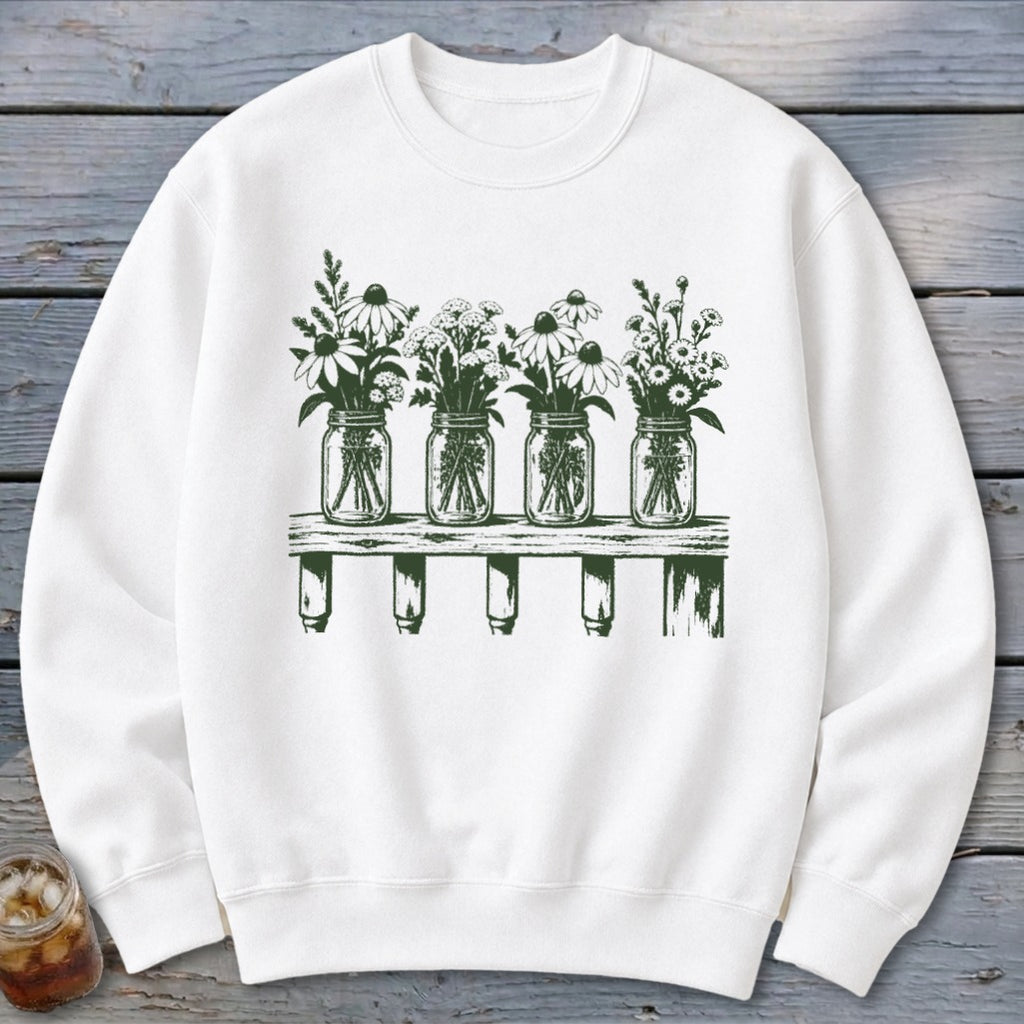 A white crewneck sweatshirt featuring a green graphic of four mason jars filled with wildflowers on a wooden shelf.