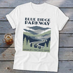 A white short-sleeved t-shirt features a Blue Ridge Parkway graphic with mountains, a winding road, and evergreen trees.