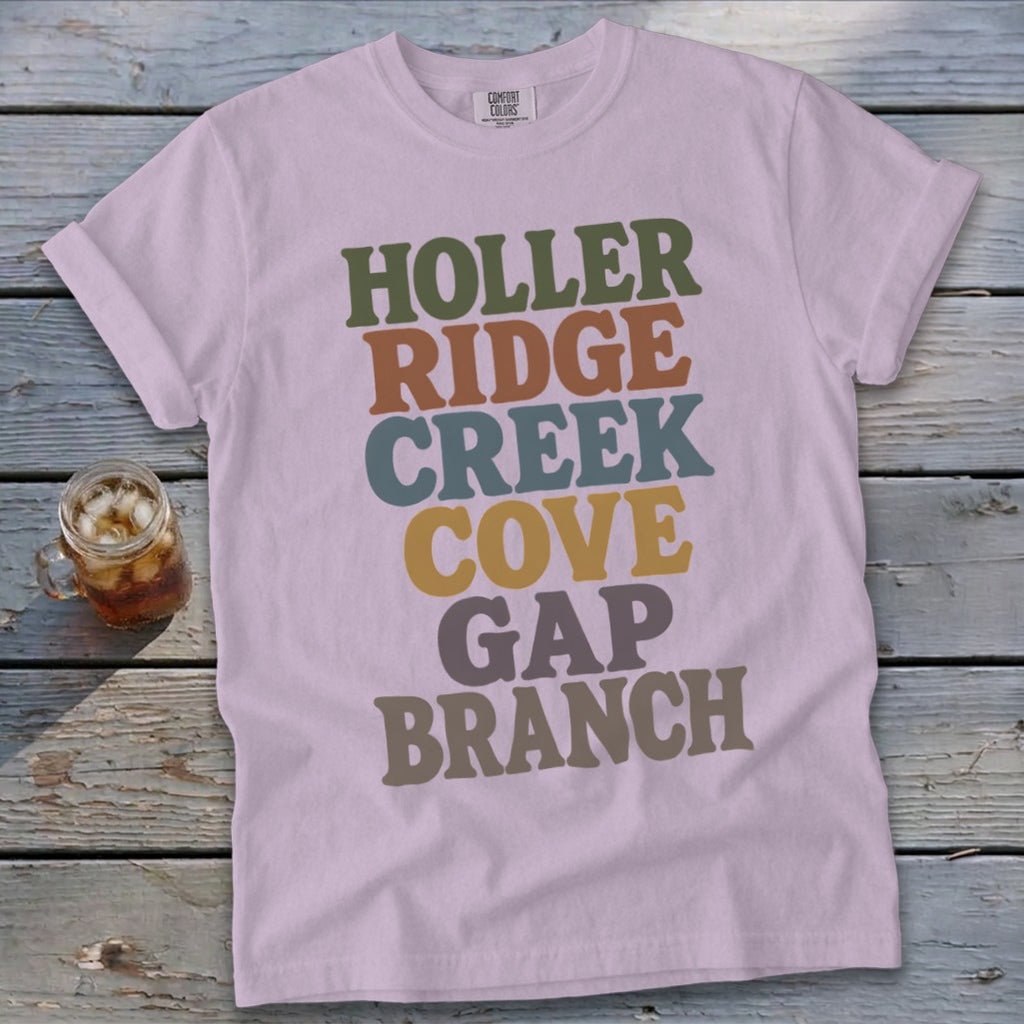 Light purple t-shirt featuring stacked multicolored retro text resting on a weathered wooden surface next to a glass of iced tea.