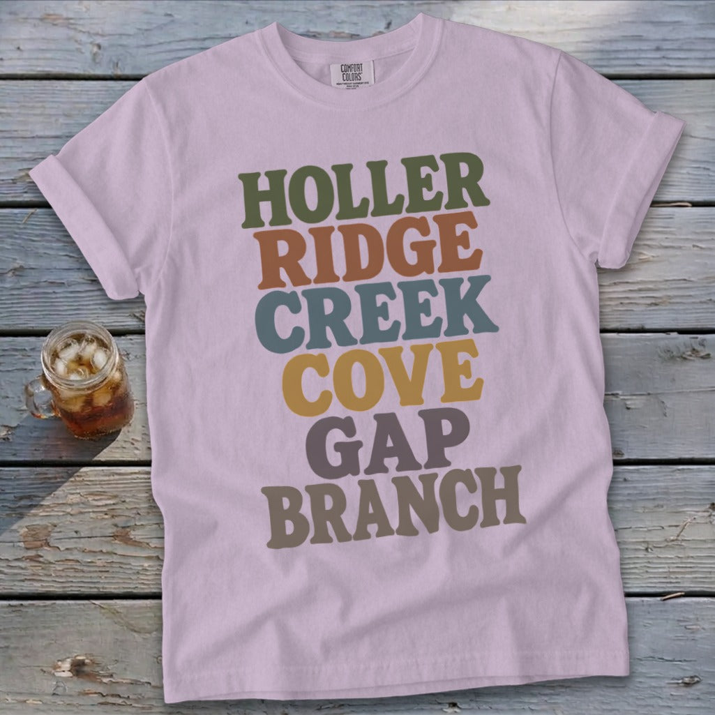 Light purple t-shirt featuring stacked multicolored retro text resting on a weathered wooden surface next to a glass of iced tea.