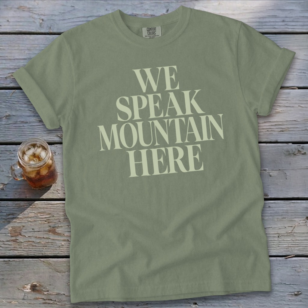 Olive green t-shirt with text "WE SPEAK MOUNTAIN HERE" and a glass of iced tea.