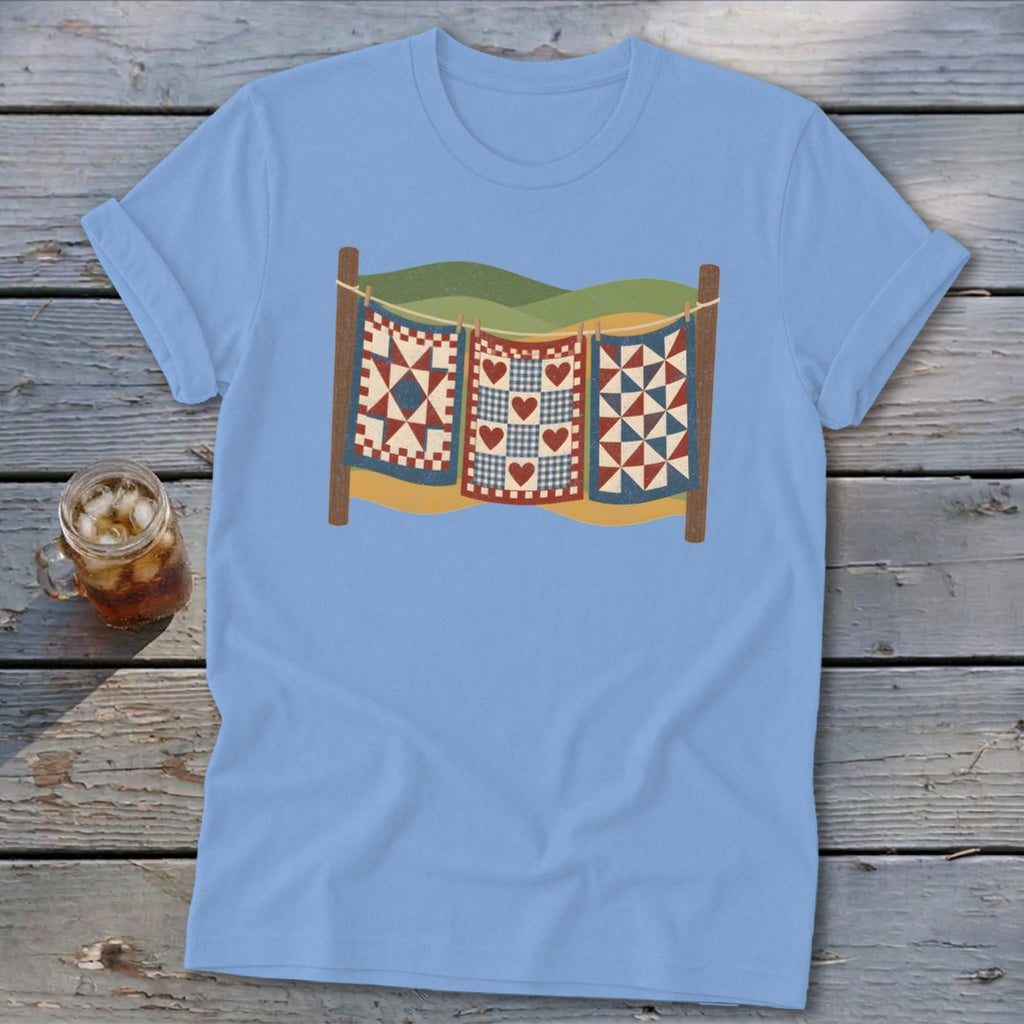 Light blue t-shirt featuring a graphic of three patchwork quilts hanging on a clothesline outdoors.