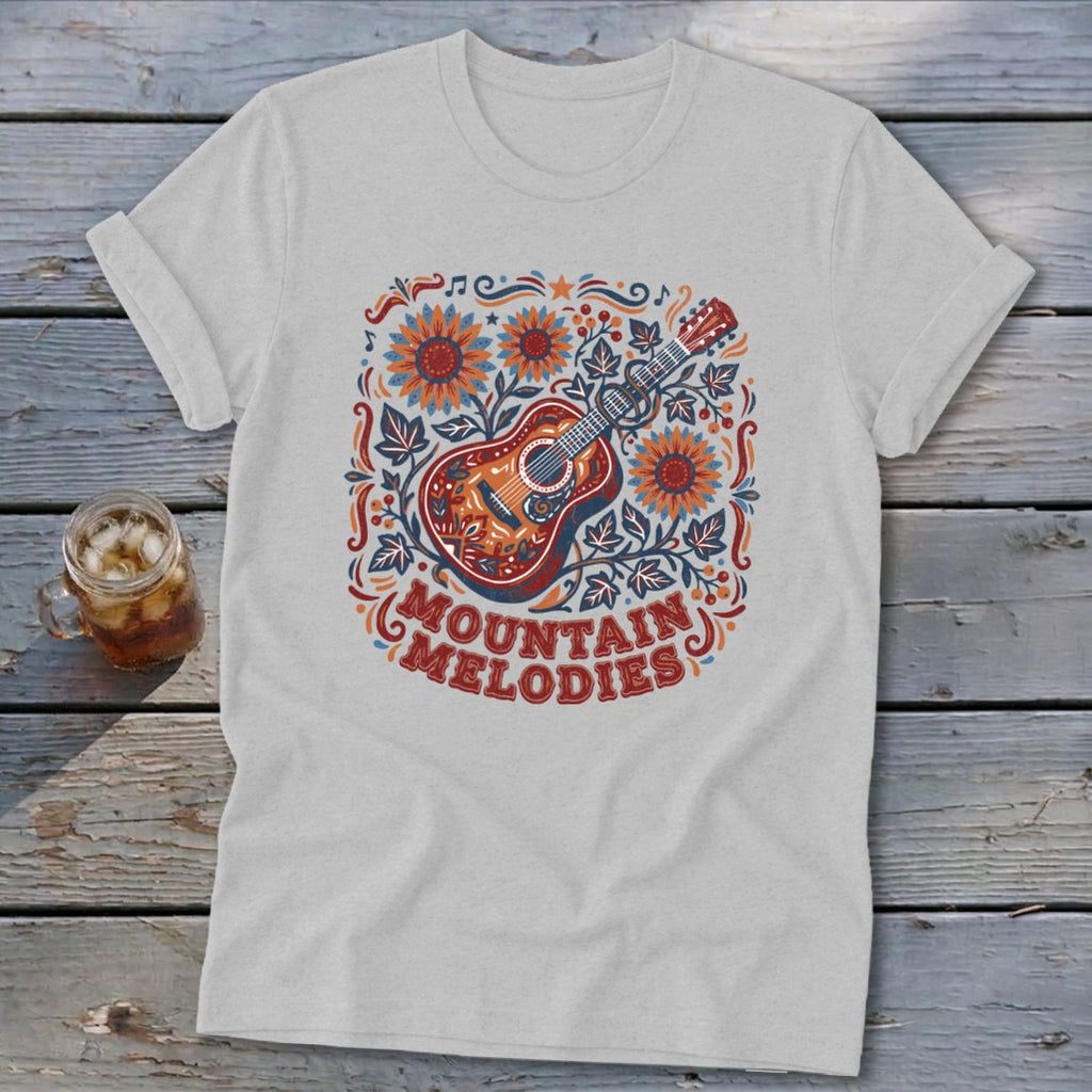 A light grey t-shirt featuring a colorful folk art graphic of a guitar, sunflowers, and the text MOUNTAIN MELODIES.