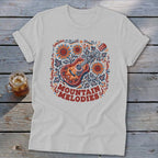 A light grey t-shirt featuring a colorful folk art graphic of a guitar, sunflowers, and the text MOUNTAIN MELODIES.
