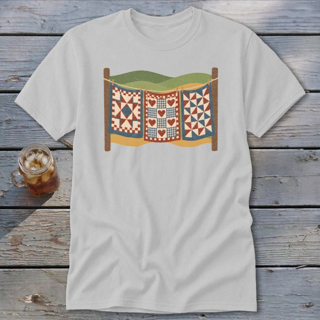 A light grey t-shirt featuring a graphic of three colorful quilts hanging on a clothesline outdoors.