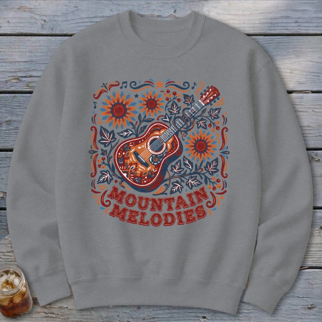 A gray crewneck sweatshirt features a folk art guitar graphic surrounded by sunflowers and the text "MOUNTAIN MELODIES".
