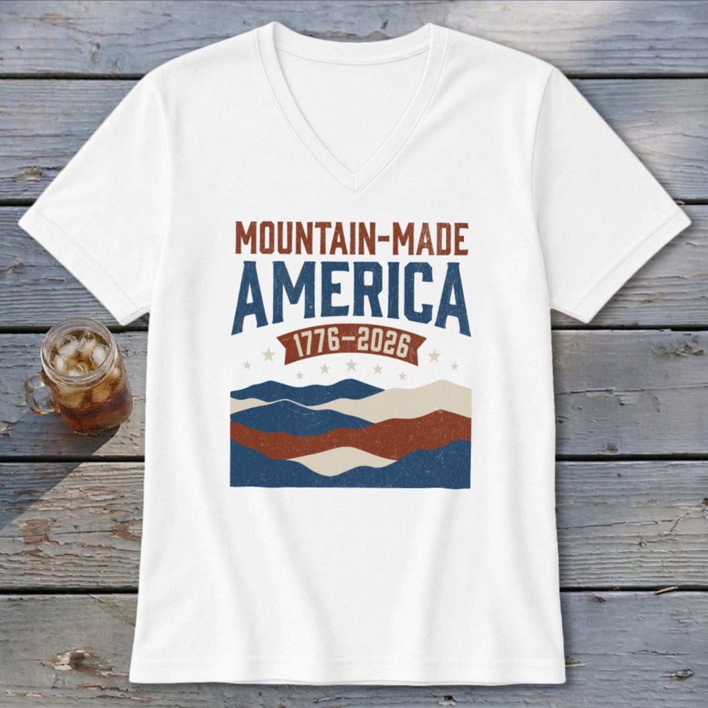 A white V-neck t-shirt featuring Mountain-Made America 1776-2026 graphic with mountains, stars, and stripes.