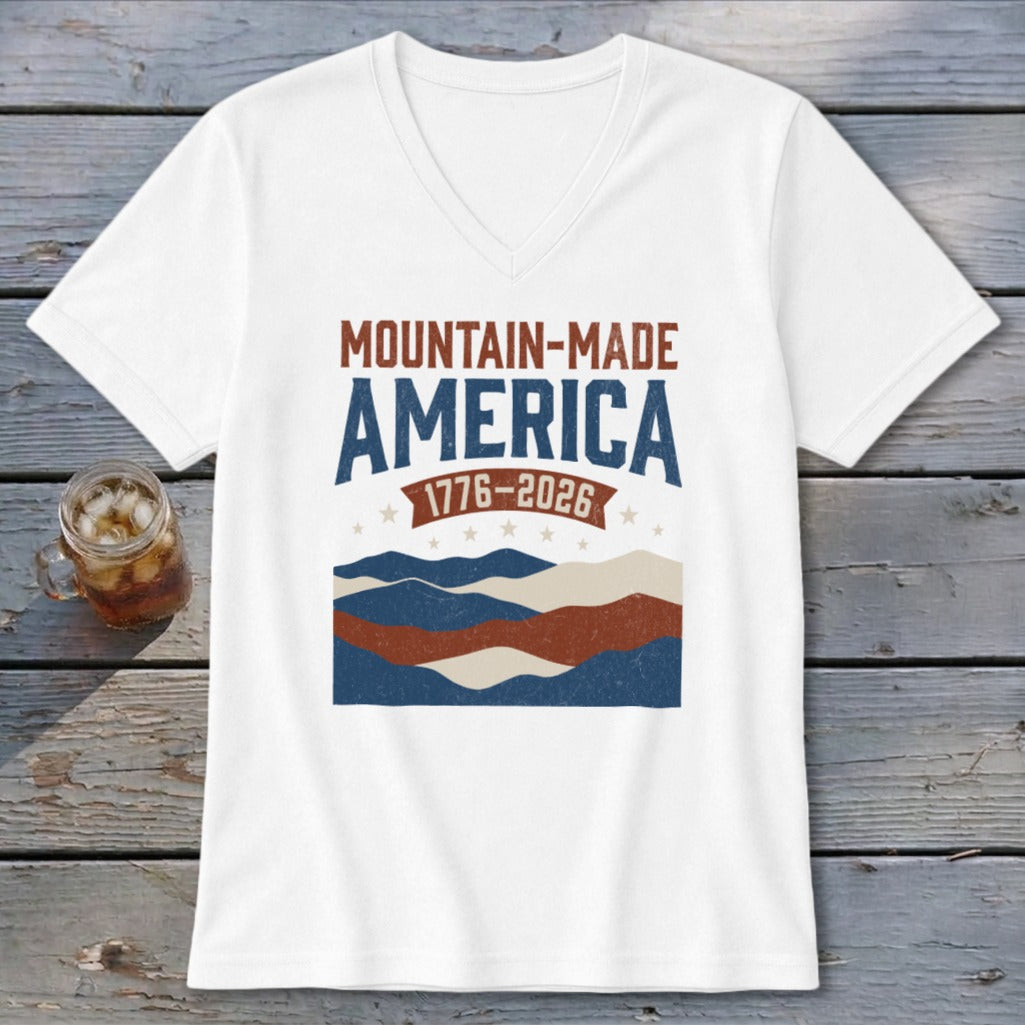 A white V-neck t-shirt featuring Mountain-Made America 1776-2026 graphic with mountains, stars, and stripes.