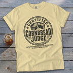 Light yellow t-shirt with a black round graphic reading Certified Cornbread Judge and a small drink.