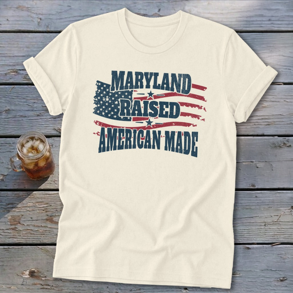 A cream-colored t-shirt on a wooden surface displays the text Maryland Raised American Made over a distressed American flag graphic.