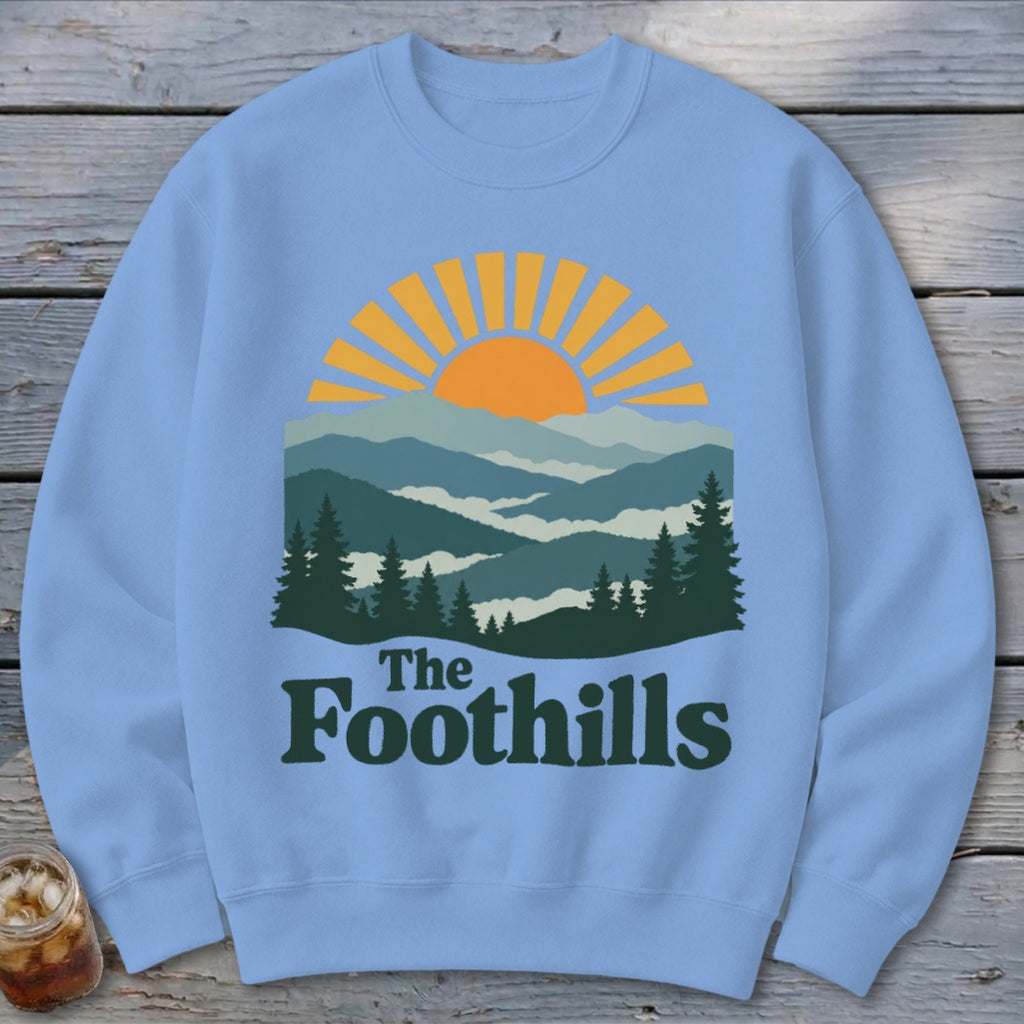 Light blue sweatshirt with a graphic of a sunrise over mountains and "The Foothills" text.