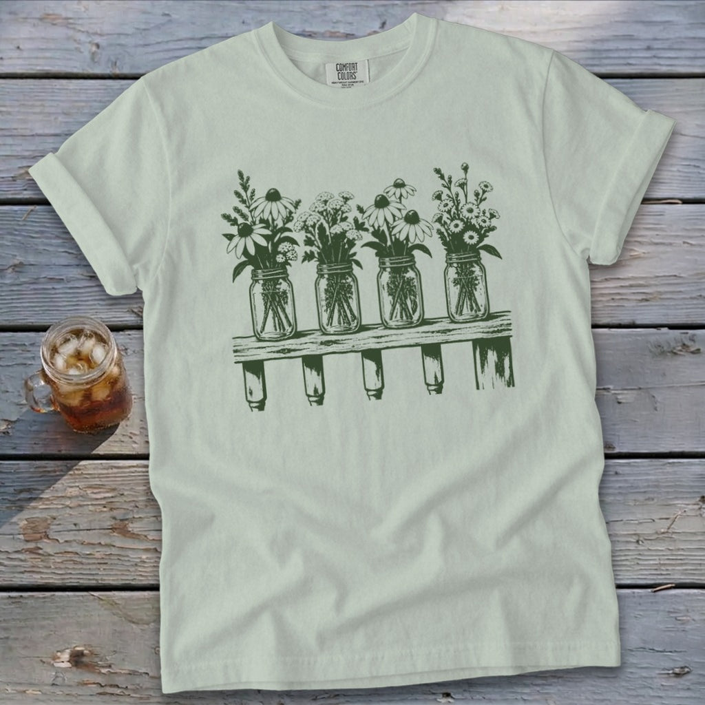 A sage green crewneck t-shirt featuring a dark green graphic of four mason jars filled with wildflowers resting on a wooden shelf.