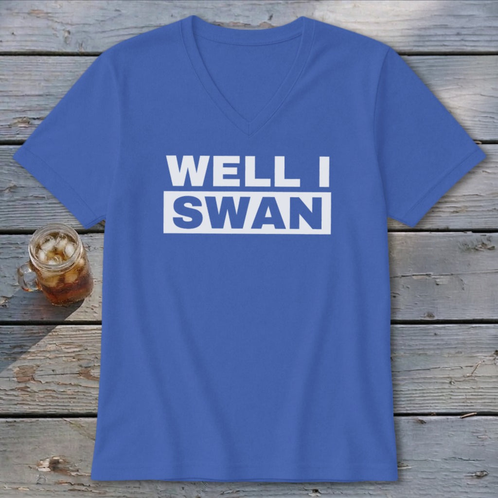 Blue V-neck t-shirt with the white text WELL I SWAN printed on the chest, lying on a rustic wooden table next to a glass of iced tea.
