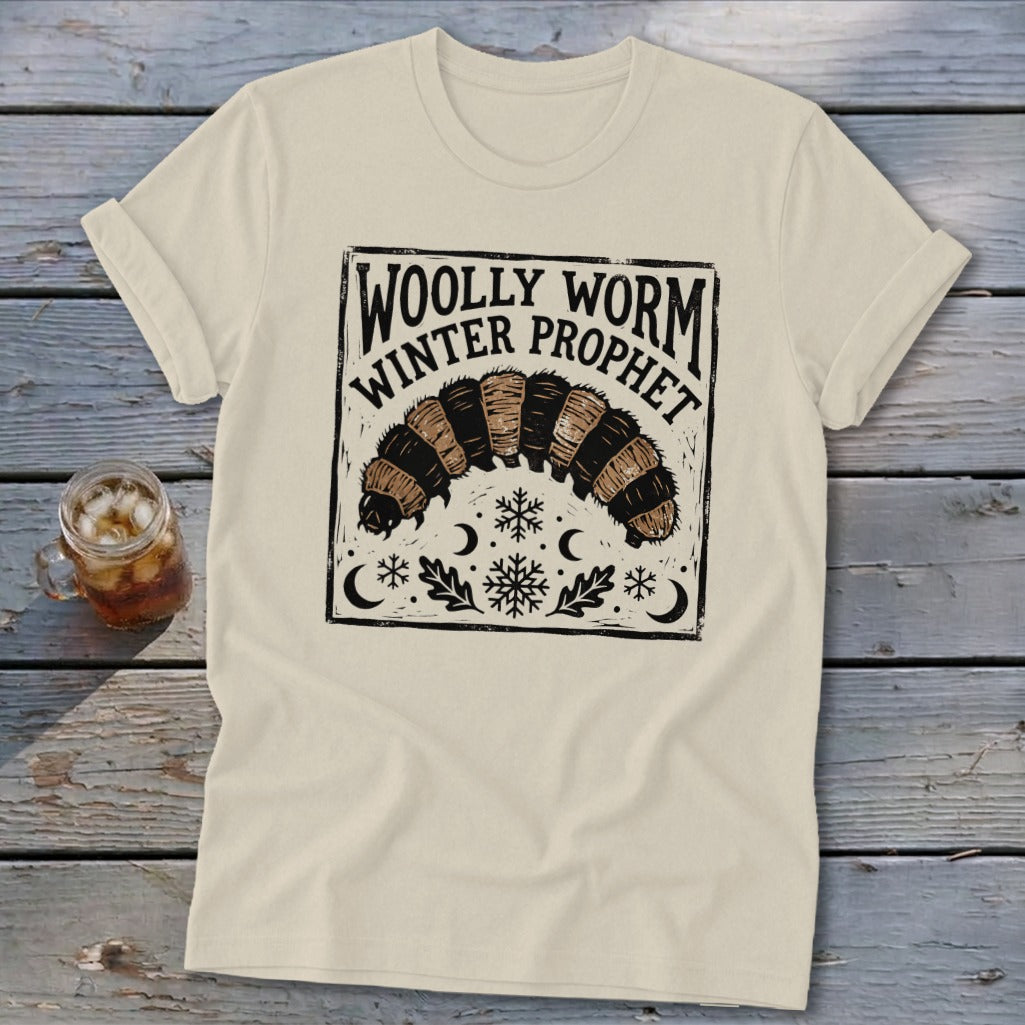 A cream-colored t-shirt features a black block-print graphic of a woolly worm, snowflakes, and the text "Woolly Worm Winter Prophet."
