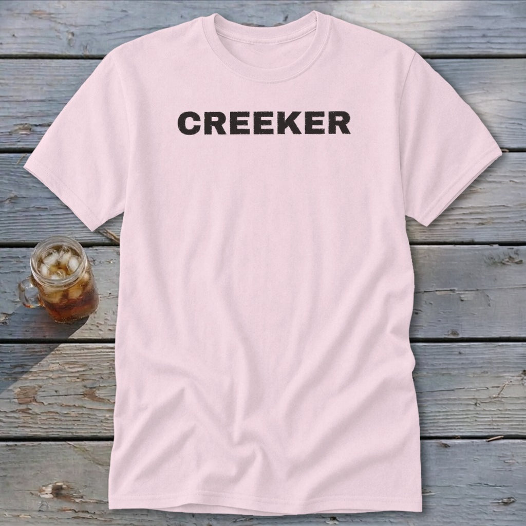 Pink t-shirt with "CREEKER" printed in black text, next to a glass of iced tea.