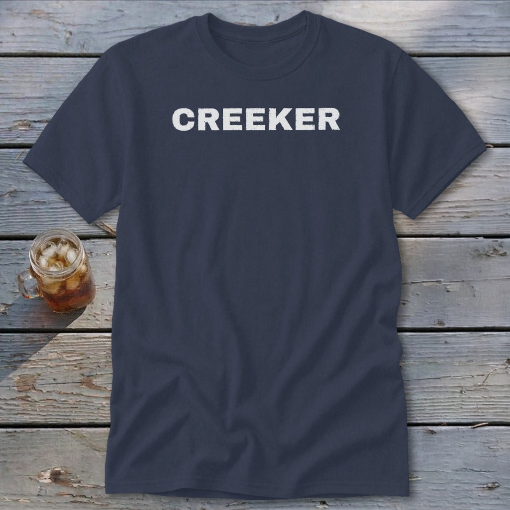 Navy t-shirt with "CREEKER" in white distressed letters, on a wooden surface with a drink.