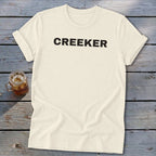 Cream t-shirt with "CREEKER" text in black, next to a glass of iced beverage.