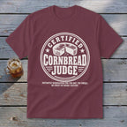Maroon short-sleeved t-shirt featuring a white round graphic with a cornbread illustration and text.