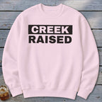 Light pink crewneck sweatshirt with black text "CREEK RAISED" across the chest.