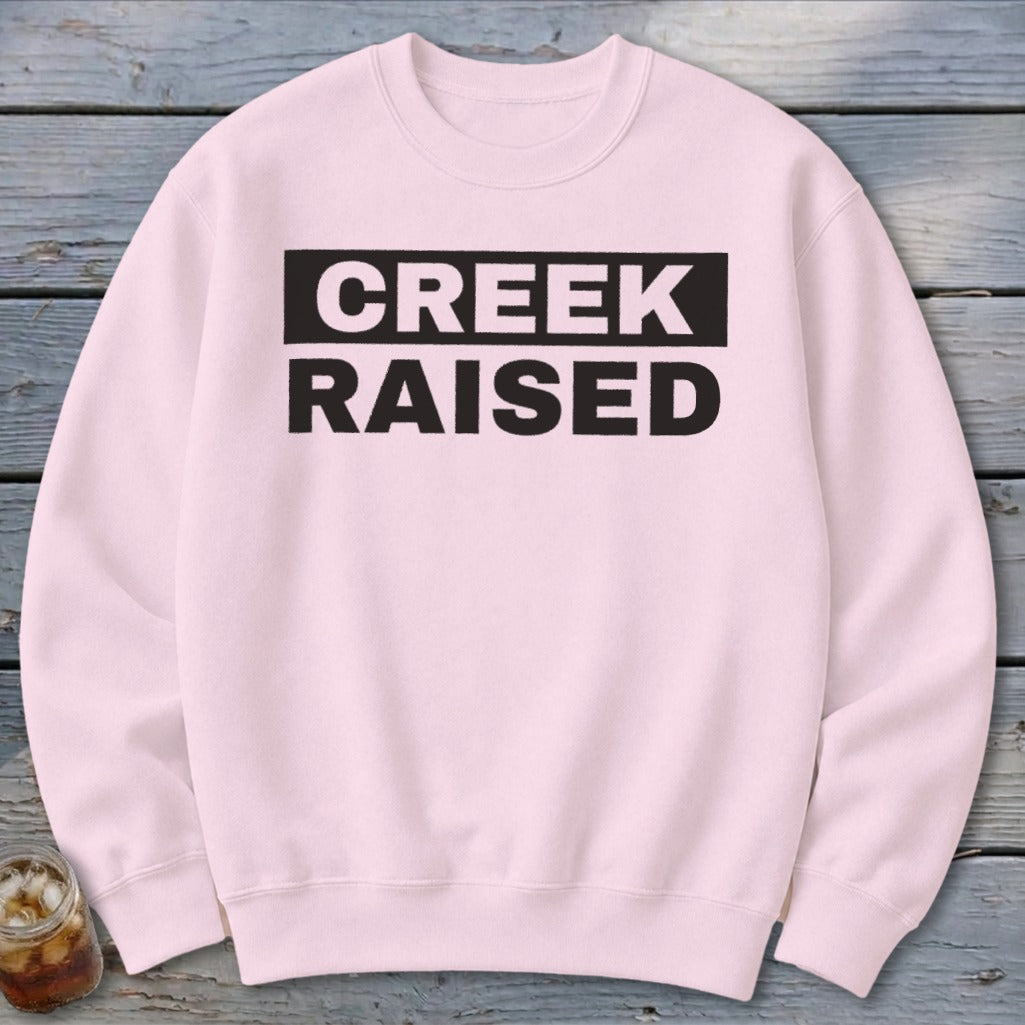 Light pink crewneck sweatshirt with black text "CREEK RAISED" across the chest.