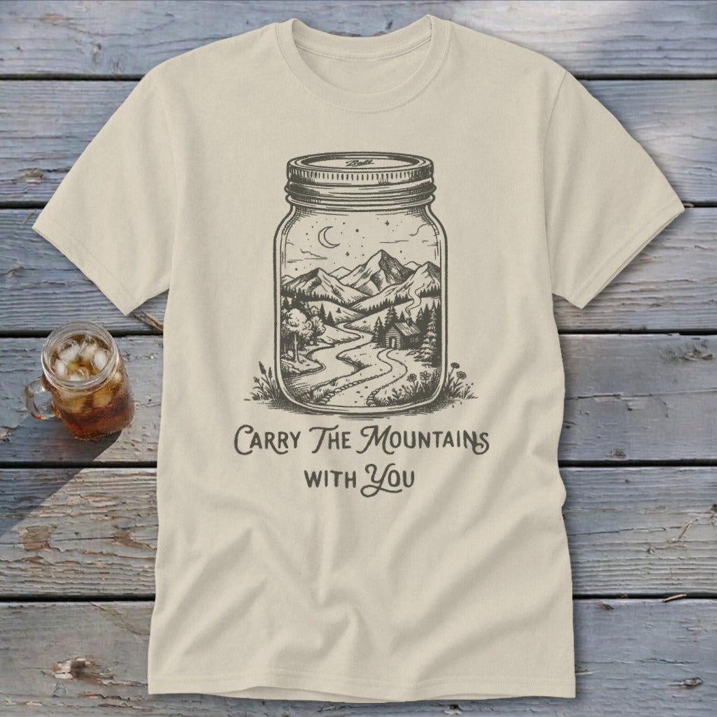 A cream-colored t-shirt featuring a black line art graphic of a landscape inside a mason jar, with the text "Carry The Mountains With You" below the jar.
