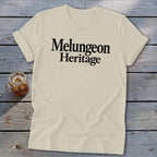 A cream-colored t-shirt with rolled sleeves lies on a rustic wooden background, featuring black text that reads "Melungeon Heritage."