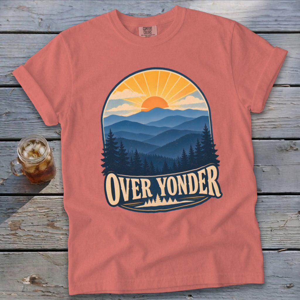 Coral t-shirt featuring a mountain sunset graphic with the text Over Yonder, lying on a wooden surface.