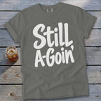 Grey t-shirt with white text reading Still A-Goin' on a weathered wood background.