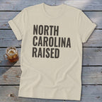 Cream t-shirt with bold black text reading "NORTH CAROLINA RAISED."