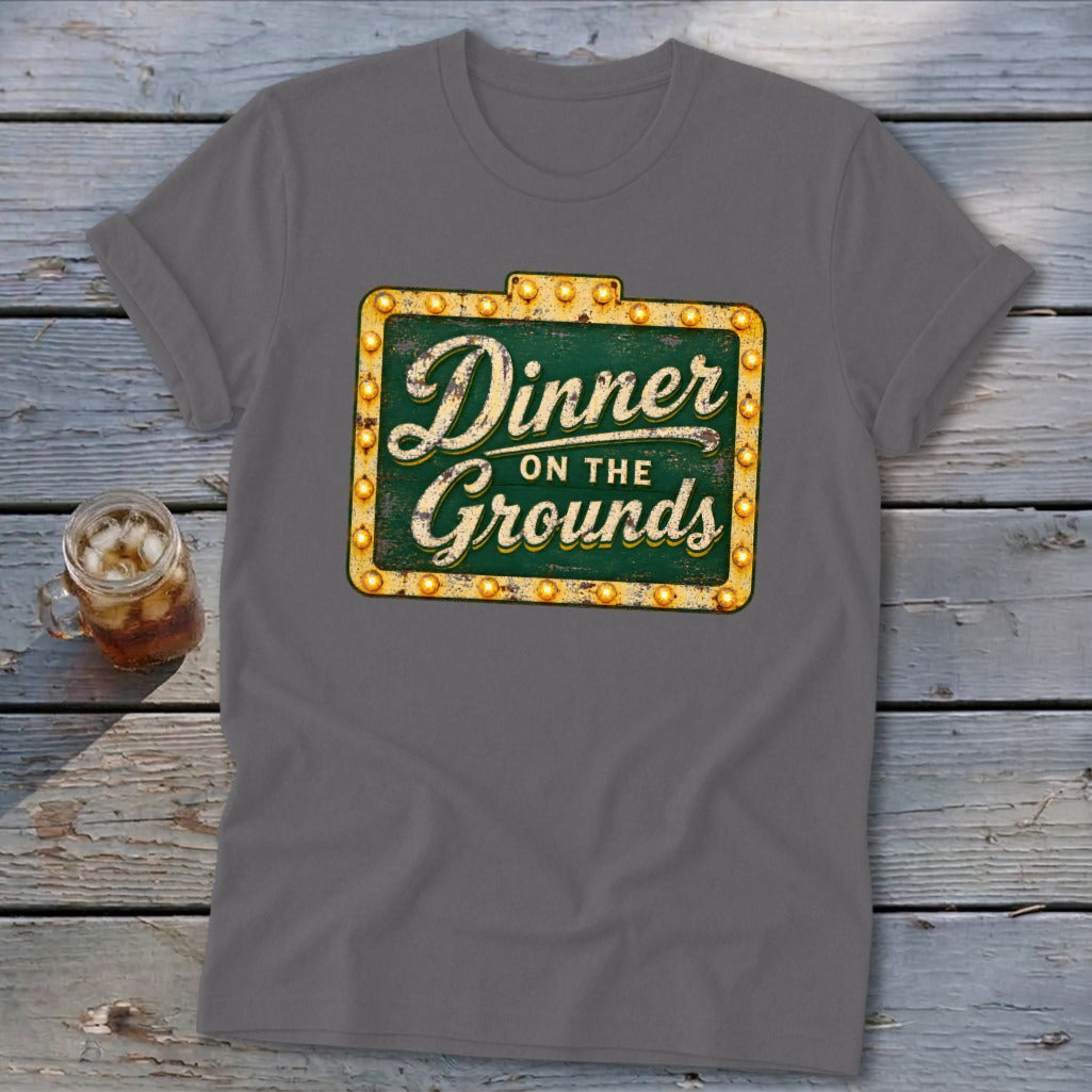 A dark gray t-shirt featuring a green vintage-style sign graphic that reads Dinner on the Grounds.