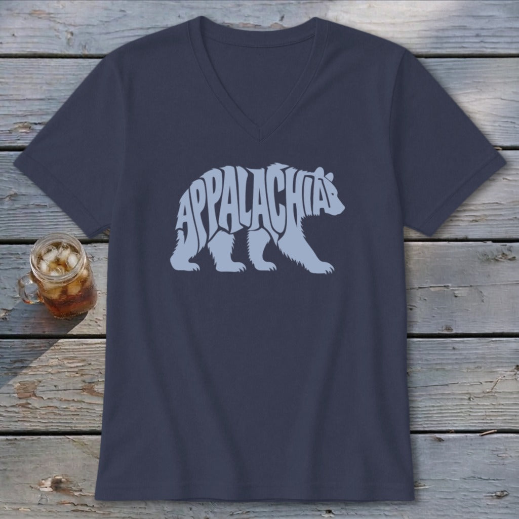 A dark blue V-neck T-shirt featuring a light blue graphic of a walking bear with the text Appalachia formed inside its body, laid flat on a rustic wooden surface next to a glass of iced tea.