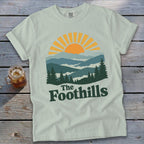 Light green t-shirt with "The Foothills" text and mountain sunset graphic.