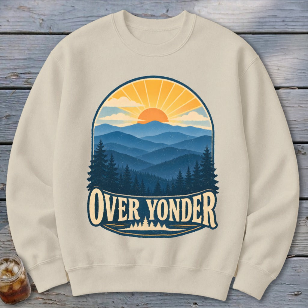 A cream-colored crewneck sweatshirt featuring a graphic of mountains, pine trees, and a setting sun with the text Over Yonder.