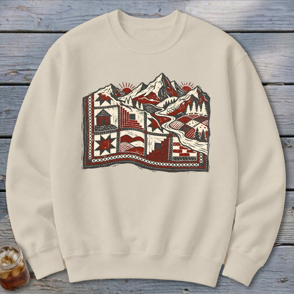 Beige crewneck sweatshirt featuring a graphic of a quilted landscape with mountains, cabins, and pine trees.