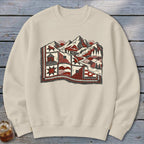 Beige crewneck sweatshirt featuring a graphic of a quilted landscape with mountains, cabins, and pine trees.