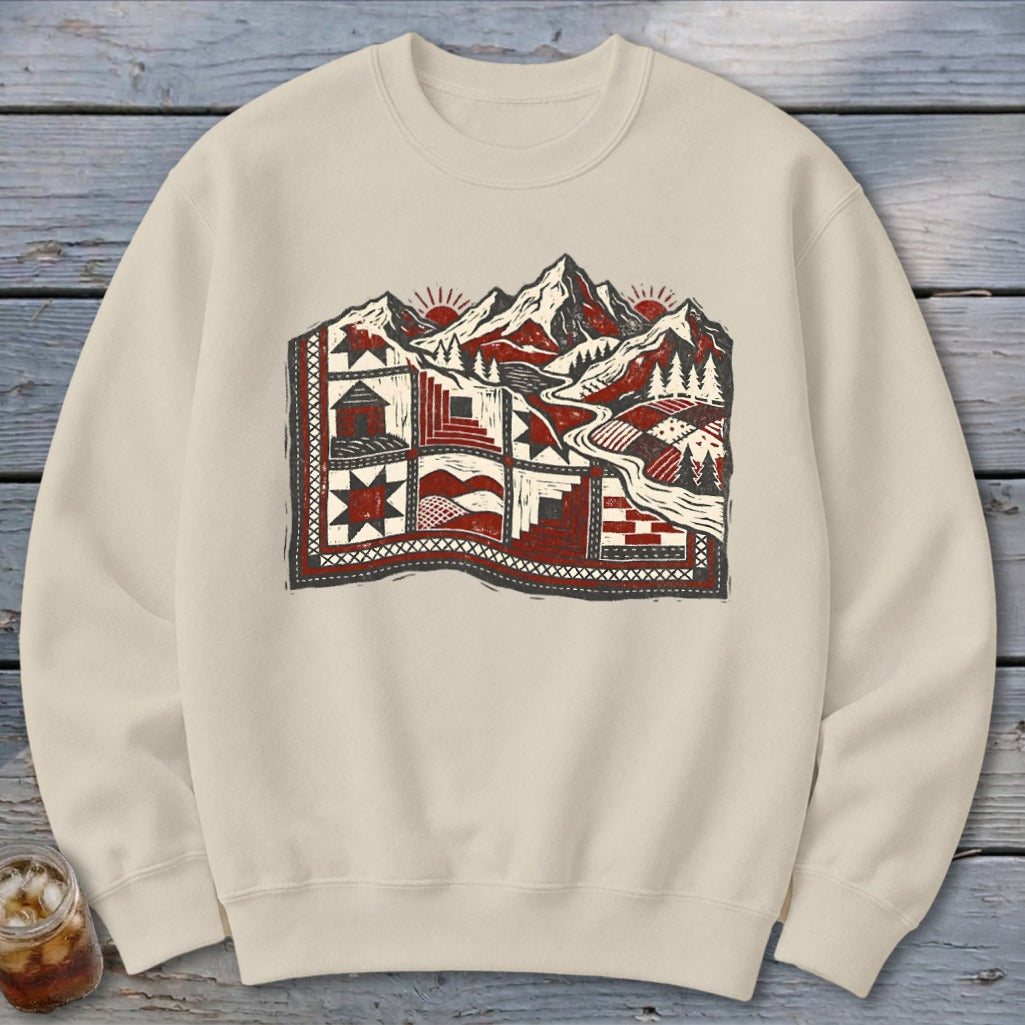 Beige crewneck sweatshirt featuring a graphic of a quilted landscape with mountains, cabins, and pine trees.